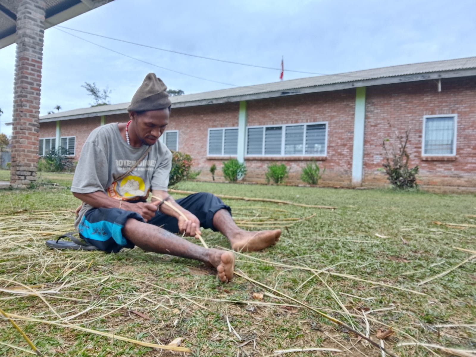 Seorang anak muda sedang mengikis dan membersihkan rotan sebagai bagian dari persiapan bahan untuk mengikat pelepah sagu kering yang digunakan sebagai atap rumah adat. Kegiatan ini merupakan salah satu tahapan dalam proses pembangunan rumah adat dengan memanfaatkan bahan dan keterampilan tradisional.